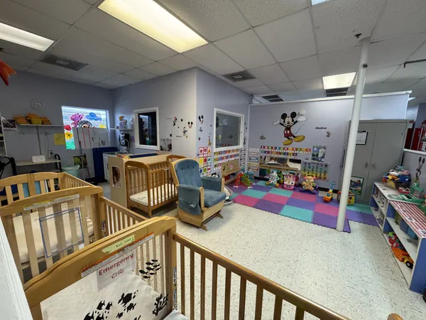 Infant room with wooden cribs, a blue glider chair, and a colorful foam play mat
