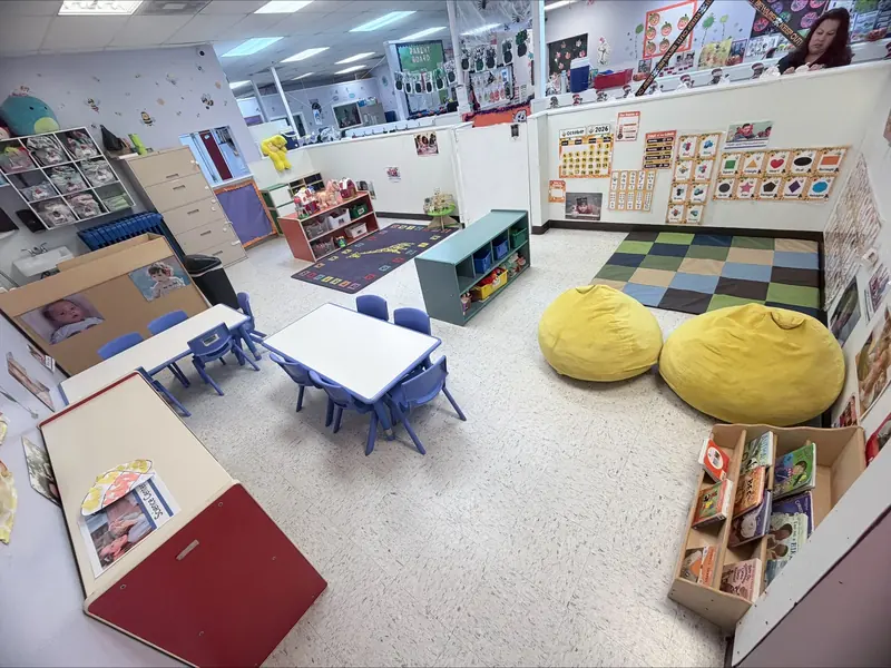 Toddler classroom with small tables, blue chairs, yellow bean bags, and a reading corner